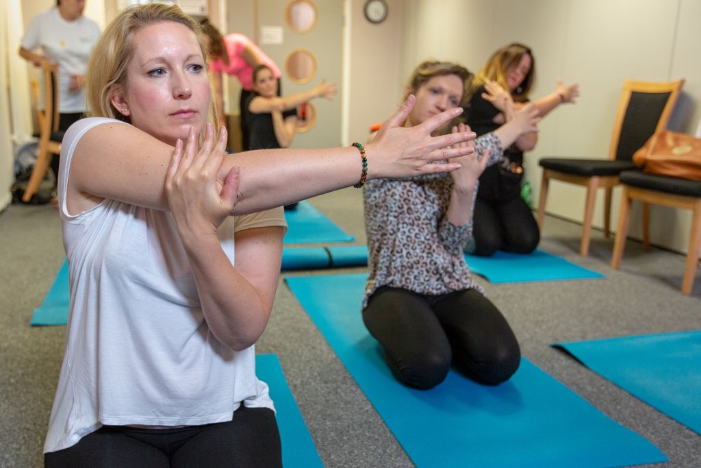 Yoga class with women in kneeling, stretched-arm positions on blue yoga mats.