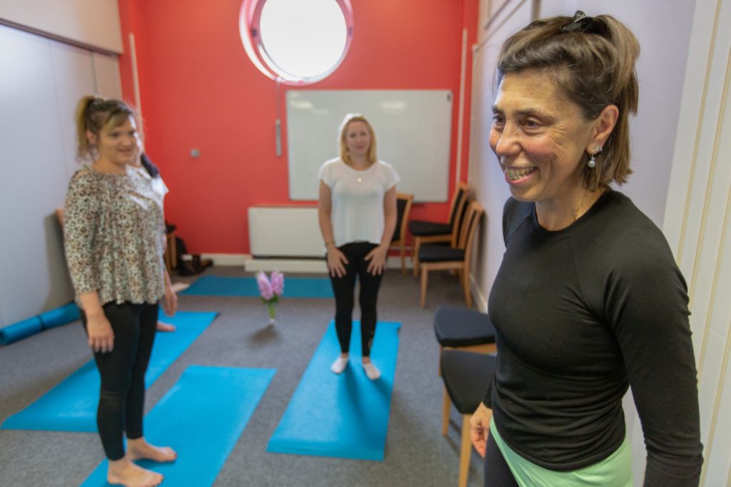 Yoga class with instructor. Women are standing on blue yoga mats and there is a vase of flowers in the centre of the floor.